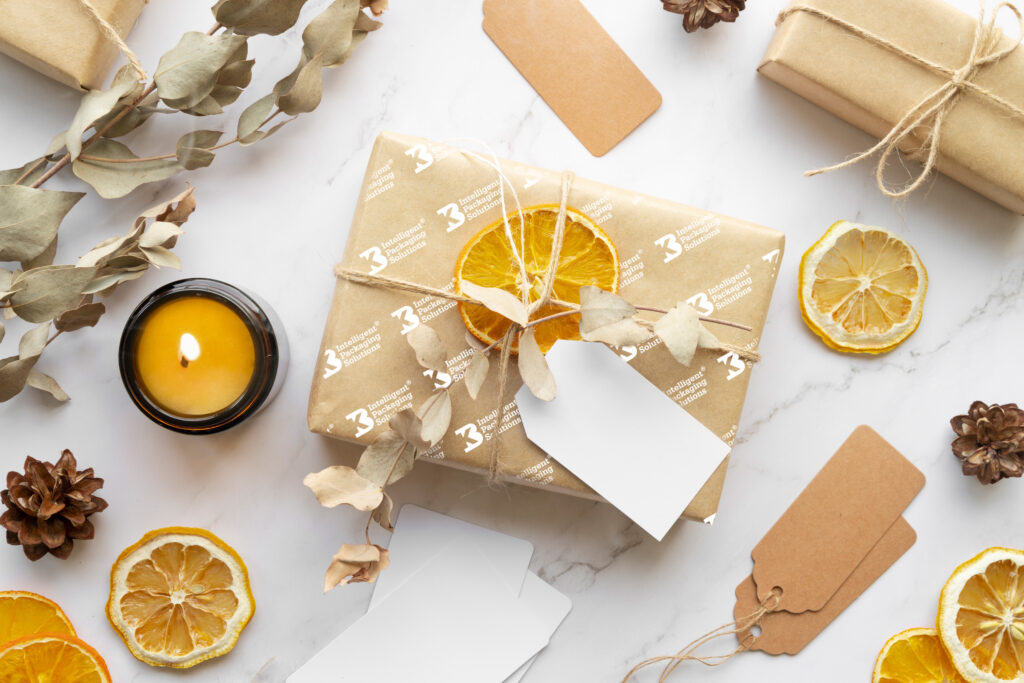 Flat lay of eco-friendly gift wrapping with brown kraft paper, twine, and dried orange slices on a white marble surface. Includes a lit yellow candle in a black holder, pinecones, eucalyptus branches, blank gift tags, and additional dried citrus slices arranged decoratively.