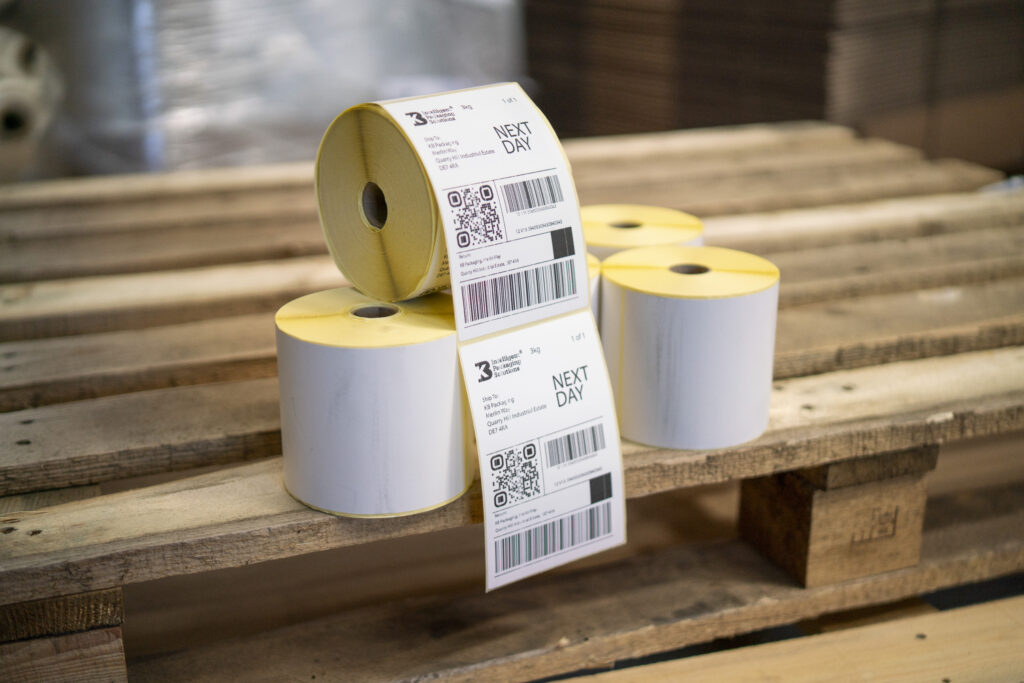 Rolls of thermal shipping labels displayed on wooden pallets in a warehouse, with printed barcode and next-day delivery labels visible.