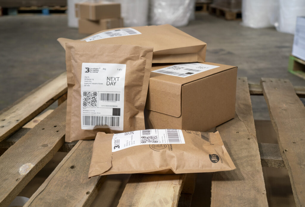 Mailing bags and cardboard boxes with printed thermal shipping labels displayed on wooden pallets in a warehouse environment.